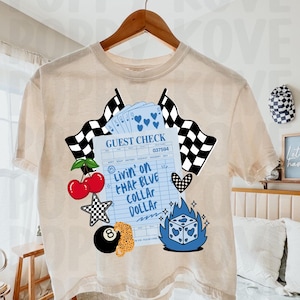 May include: Cream-colored t-shirt with a graphic design featuring a blue guest check with the text "Livin' on that Blue Collar Dollar." The design includes cherries, dice, playing cards, and checkered flags. The shirt is hanging on a wooden hanger.
