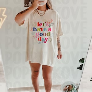 May include: A white oversized t-shirt with a colorful graphic that says "Let's have a good day" surrounded by school supplies.