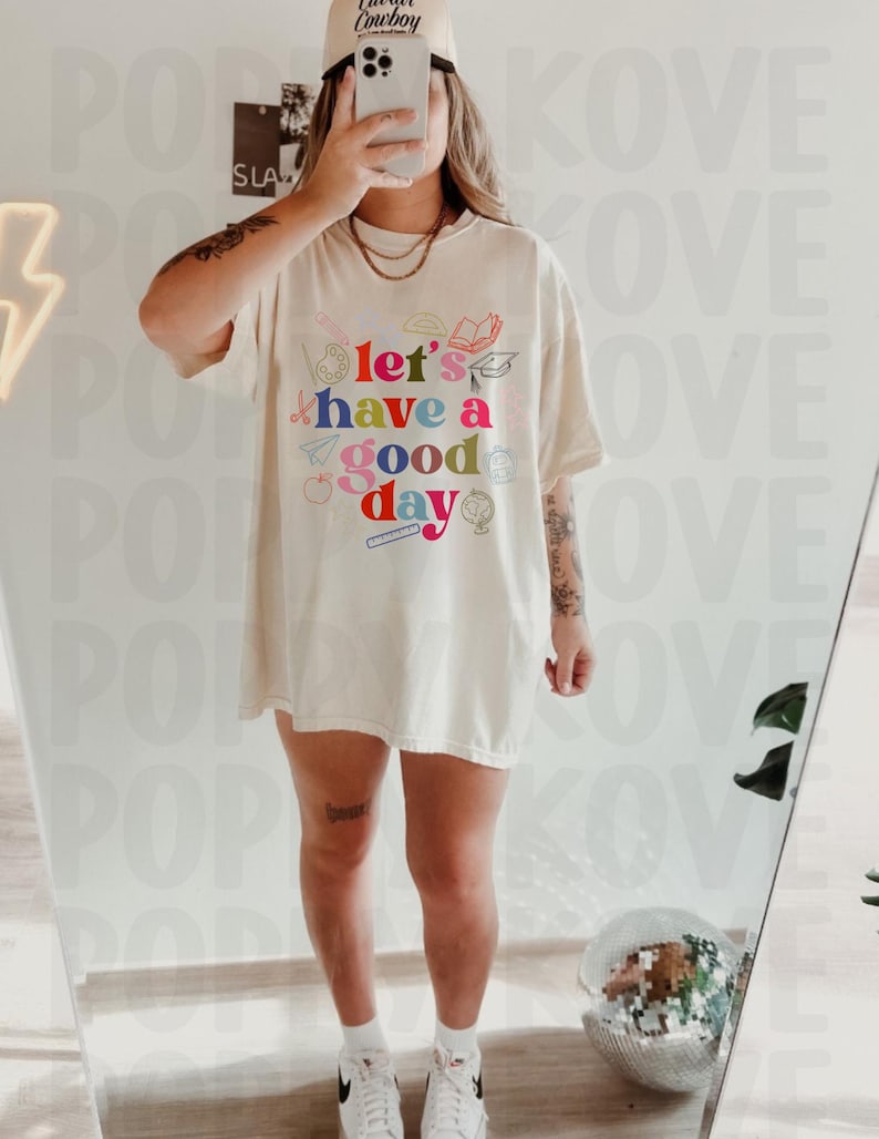 May include: A white oversized t-shirt with a colorful graphic that says "Let's have a good day" surrounded by school supplies.