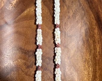 Könnte beinhalten: Eine weiße Muschel-Lei mit braunen Akzenten. Die Lei besteht aus kleinen, weißen Muscheln, die auf einer Schnur zusammengefügt sind. Die Lei ist etwa 60 cm lang.