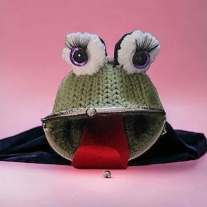 May include: A quirky, handmade frog-shaped purse. It features a knitted green body, large googly eyes with long eyelashes, and a red felt tongue. The purse has a silver clasp and is set against a pink backdrop.