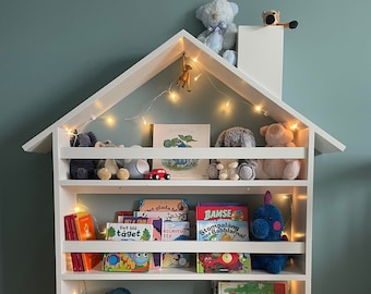 May include: White wooden bookshelf shaped like a house with five shelves. The shelves are filled with children's books and stuffed animals. There are fairy lights strung across the top of the bookshelf.