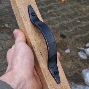 Könnte beinhalten: Ein schwarzer, gebogener Metallgriff ist mit zwei schwarzen Schrauben an einem Holzbrett befestigt. Der Griff hat eine leicht strukturierte Oberfläche. Das Holz ist hellbraun. Der Griff ist zum Ziehen oder Greifen bestimmt.