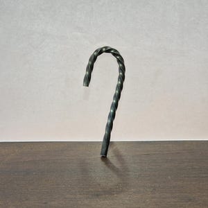 May include: A hand-forged metal candy cane with a twisted design. The dark gray cane stands upright on a dark wood surface against a light gray background. The cane's hook shape is a classic holiday symbol.