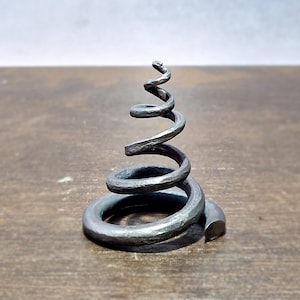 Może przedstawiać: Ręcznie wykonana, ciemnoszara metalowa rzeźba w kształcie spirali, przypominająca stylizowane drzewo lub element dekoracyjny. Rzeźba umieszczona jest na drewnianej powierzchni, ukazując jej teksturowany, kuty wygląd.