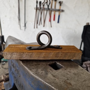 Peut inclure: Un crochet en métal forgé à la main avec un dessus en boucle, monté sur une base en bois rectangulaire. Le crochet est gris foncé et le bois est brun chaud. L'arrière-plan montre des outils de forgeron accrochés à un mur blanc. Le crochet est probablement pour suspendre des objets.
