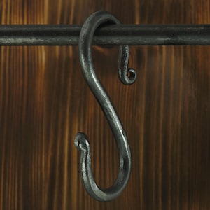 Peut inclure: Un crochet en S en métal gris foncé forgé à la main, suspendu à une tige assortie. Le crochet a une surface texturée et une extrémité recourbée. L'arrière-plan est une surface en bois avec un grain vertical.