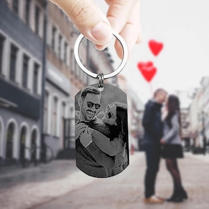May include: A silver metal keychain with a rectangular pendant. The pendant has a black and white engraved image of a couple embracing. The keychain is held up by a hand, with a blurred street scene in the background.