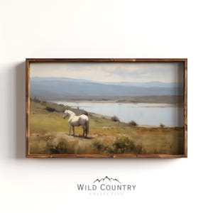 May include: A framed landscape painting depicts a white horse standing on a grassy hill overlooking a lake and distant mountains. The artwork features a rustic wooden frame and a serene, natural color palette.