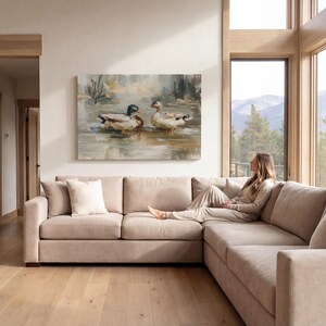 A woman sits on a beige sectional sofa in a spacious living room, gazing out a large window at a serene lake and mountains in the distance.