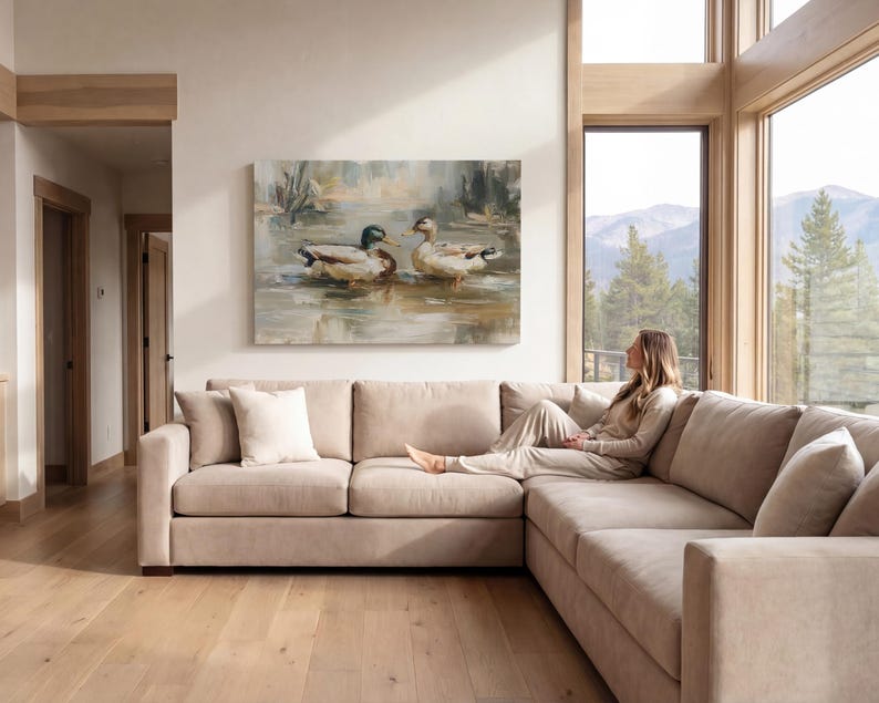 A woman sits on a beige sectional sofa in a spacious living room, gazing out a large window at a serene lake and mountains in the distance.