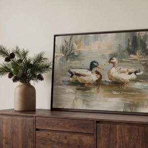 a wooden cabinet with a framed painting of two ducks swimming in a pond, placed on top of it. Next to the painting, there is a vase with pine branches and pine cones.