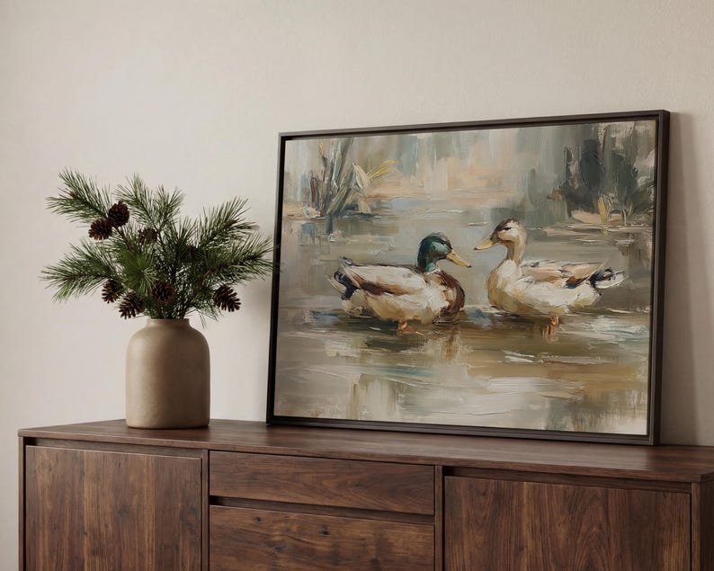 a wooden cabinet with a framed painting of two ducks swimming in a pond, placed on top of it. Next to the painting, there is a vase with pine branches and pine cones.