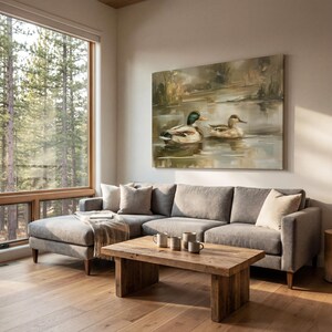 a cozy living room with a large window overlooking a wooded area, allowing natural light to fill the space. The focal point is a comfortable gray sofa with a wooden coffee table in front of it, and a large painting of ducks swimming in a pond hanging on the wall above the sofa.