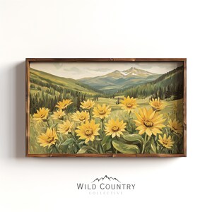 Puede incluir: Una impresión artística rectangular enmarcada que muestra un paisaje de montañas detrás de un campo de girasoles amarillos. La escena incluye colinas verdes, pinos y un cielo claro. Debajo del marco aparece el texto Wild Country Collective.