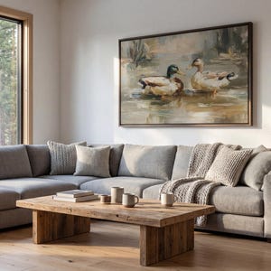 a cozy living room with a large painting of ducks on the wall, a gray sectional sofa, a wooden coffee table, and a large window overlooking a wooded area.