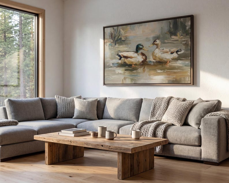 a cozy living room with a large painting of ducks on the wall, a gray sectional sofa, a wooden coffee table, and a large window overlooking a wooded area.