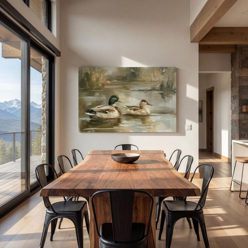 a dining room with a large wooden table surrounded by black metal chairs. A painting of ducks swimming in a pond is displayed on the wall above the table. The room has large windows that provide a scenic view of the outdoors, and the overall decor has a modern and minimalist aesthetic.