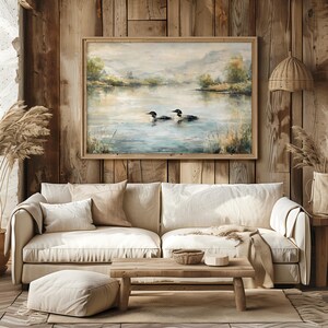 a cozy living room with a large painting of ducks swimming on a lake hanging on the wall above a beige sofa. The room has a rustic, cabin-like atmosphere with wooden walls and a wooden floor.