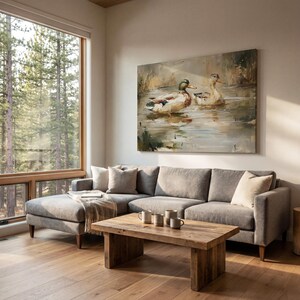 a cozy living room with a large window overlooking a wooded area, allowing natural light to fill the space. The focal point is a comfortable gray sofa adorned with throw pillows, and a wooden coffee table in front of it. Above the sofa hangs a large painting of ducks swimming in a pond, adding a touch of nature to the room.