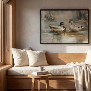 a cozy living room with a large window overlooking a scenic outdoor view. A painting of ducks swimming in a pond hangs on the wall above a wooden bench, which is adorned with white pillows and a white blanket. A small wooden table with a book and a mug is situated in front of the bench, creating a comfortable and inviting atmosphere.