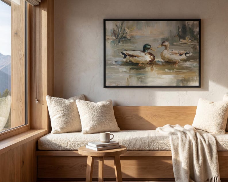 a cozy living room with a large window overlooking a scenic outdoor view. A painting of ducks swimming in a pond hangs on the wall above a wooden bench, which is adorned with white pillows and a white blanket. A small wooden table with a book and a mug is situated in front of the bench, creating a comfortable and inviting atmosphere.