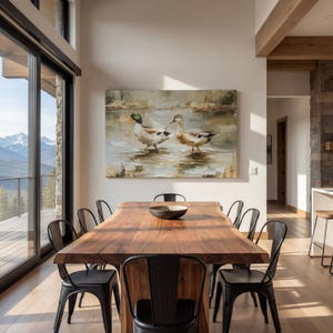 a modern dining room with a large wooden table surrounded by black metal chairs. A large painting of ducks is displayed on the wall above the table. The room has large windows that provide a scenic view of the outdoors, and the overall design has a contemporary and minimalist aesthetic.