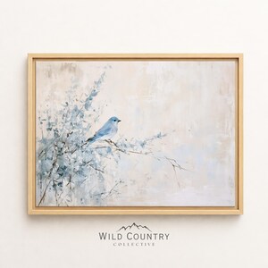 May include: A framed wall art print featuring a light blue bird perched on thin, delicate branches against a soft, neutral beige and cream background. The artwork displays a painterly texture with muted blue foliage and a light wooden frame.