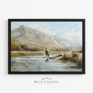 May include: A framed landscape painting depicting a person fly fishing in a river with large mountains in the background. The scene features earthy tones of brown, tan, and blue, with the text WILD COUNTRY COLLECTIVE printed below the artwork.