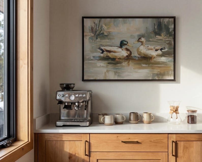 a kitchen counter with a coffee maker, coffee cups, and a framed painting of ducks swimming in a pond.