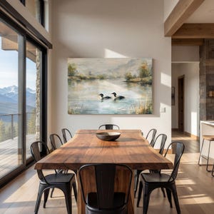a modern dining room with a large wooden table surrounded by black metal chairs. A large painting of a lake with ducks is displayed on the wall above the table. The room has large windows that provide a scenic view of the outdoors, and the overall design has a contemporary and elegant aesthetic.
