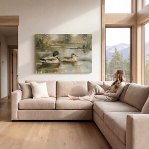 A woman sits on a beige sectional sofa in a spacious living room, gazing out a large window at a serene landscape of mountains and trees.