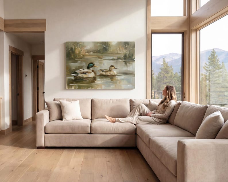 A woman sits on a beige sectional sofa in a spacious living room, gazing out a large window at a serene landscape of mountains and trees.