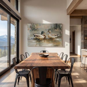 a dining room with a large wooden table surrounded by black metal chairs. Above the table, there is a painting of ducks swimming in a body of water. The room has large windows that provide a scenic view of mountains, creating a serene and picturesque atmosphere.