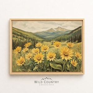 Puede incluir: Un cuadro de paisaje enmarcado que muestra un campo de flores silvestres de color amarillo brillante en primer plano, con un denso bosque de pinos verdes y montañas nevadas al fondo. La obra está en un marco de madera clara.