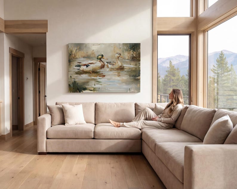 A woman sits on a beige sectional sofa in a spacious living room, gazing out a large window overlooking a scenic mountain landscape.