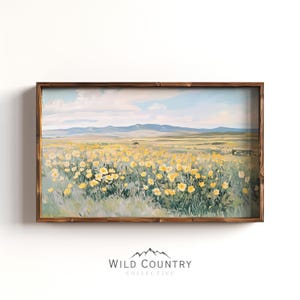 Puede incluir: Una pintura de paisaje rectangular en un marco de madera marrón oscuro que representa un campo de flores silvestres amarillas bajo un cielo azul pálido con nubes suaves y montañas distantes. Debajo del marco, el texto dice WILD COUNTRY COLLECTIVE.