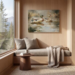 a cozy and inviting living space with a large window overlooking a serene lake, a painting of ducks swimming in the water, and a comfortable couch with a coffee mug and a book on it.