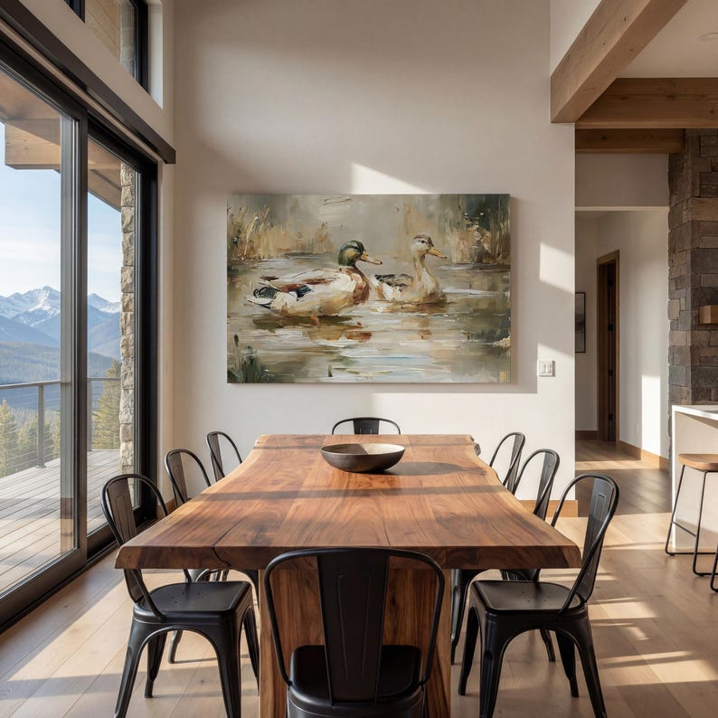 a modern dining room with a large wooden table surrounded by black metal chairs. A large painting of ducks swimming in a pond is displayed on the wall above the table. The room has large windows that provide a scenic view of the outdoors, and the overall design has a contemporary and elegant aesthetic.