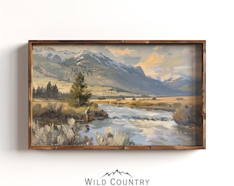 May include: A framed landscape painting depicting a person fly fishing in a river with snow-capped mountains in the background. The scene features muted tones of blue, tan, and green, with a dark wooden frame and the text Wild Country Collective.