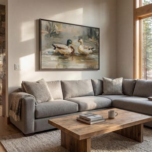 a cozy living room with a large painting of ducks on the wall, a comfortable gray sectional sofa, a wooden coffee table, and a large window overlooking a wooded area.