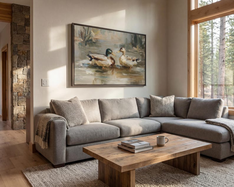 a cozy living room with a large painting of ducks on the wall, a comfortable gray sectional sofa, a wooden coffee table, and a large window overlooking a wooded area.
