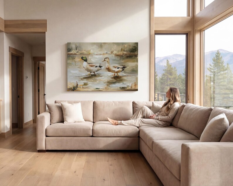A woman sits on a beige sectional sofa in a spacious living room, gazing out a large window overlooking a scenic mountain landscape.