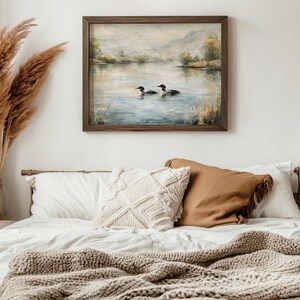 a serene bedroom scene with a framed painting of ducks swimming on a lake hanging on the wall above a bed with white pillows and a brown throw blanket.