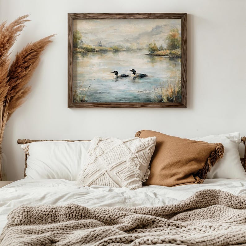 a serene bedroom scene with a framed painting of ducks swimming on a lake hanging on the wall above a bed with white pillows and a brown throw blanket.