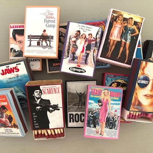 May include: A collection of matchboxes featuring movie posters. The posters include "Forrest Gump", "Clueless", "Jaws", "Scarface", "Legally Blonde", "Almost Famous", and "Rock".
