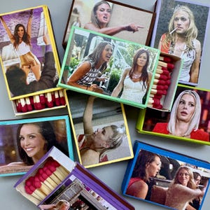 May include: A collection of decorative matchboxes, each featuring a different image of women in various poses. The matchboxes are open, revealing the red-tipped matches inside. The boxes are in various colors, including yellow, green, and blue.