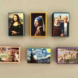 以下が含まれることがあります： 有名な絵画の小さな額装されたプリントが6つ。プリントはすべて異なる色とスタイルです。プリントはすべて明るい灰色の表面に一列に並んでいます。
