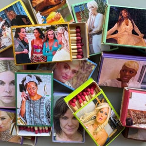 May include: A collection of matchboxes with various images of women. The matchboxes are in different colors, and some are open, revealing the red-tipped matches inside. The images on the boxes vary in style and color.
