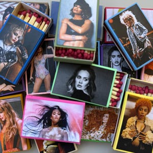 May include: A collection of matchboxes featuring various female musicians. Each box has a different color and design, with a photo of a musician on the front. The musicians include Adele, Gwen Stefani, and others.
